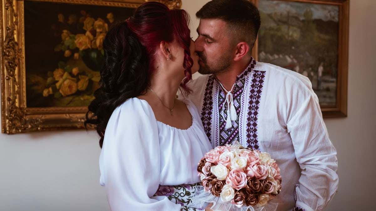 A couple in traditional Romanian attire sharing a romantic kiss during their wedding ceremony indoors.