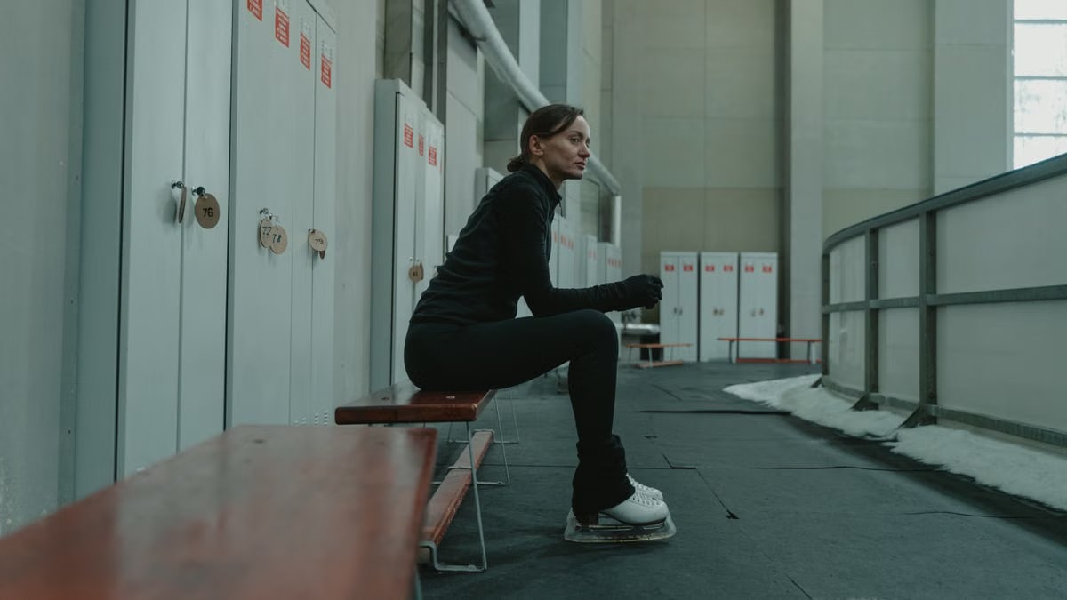 Woman sitting on a bench in an indoor ice rink, wearing ice skating gear, thoughtful expression.